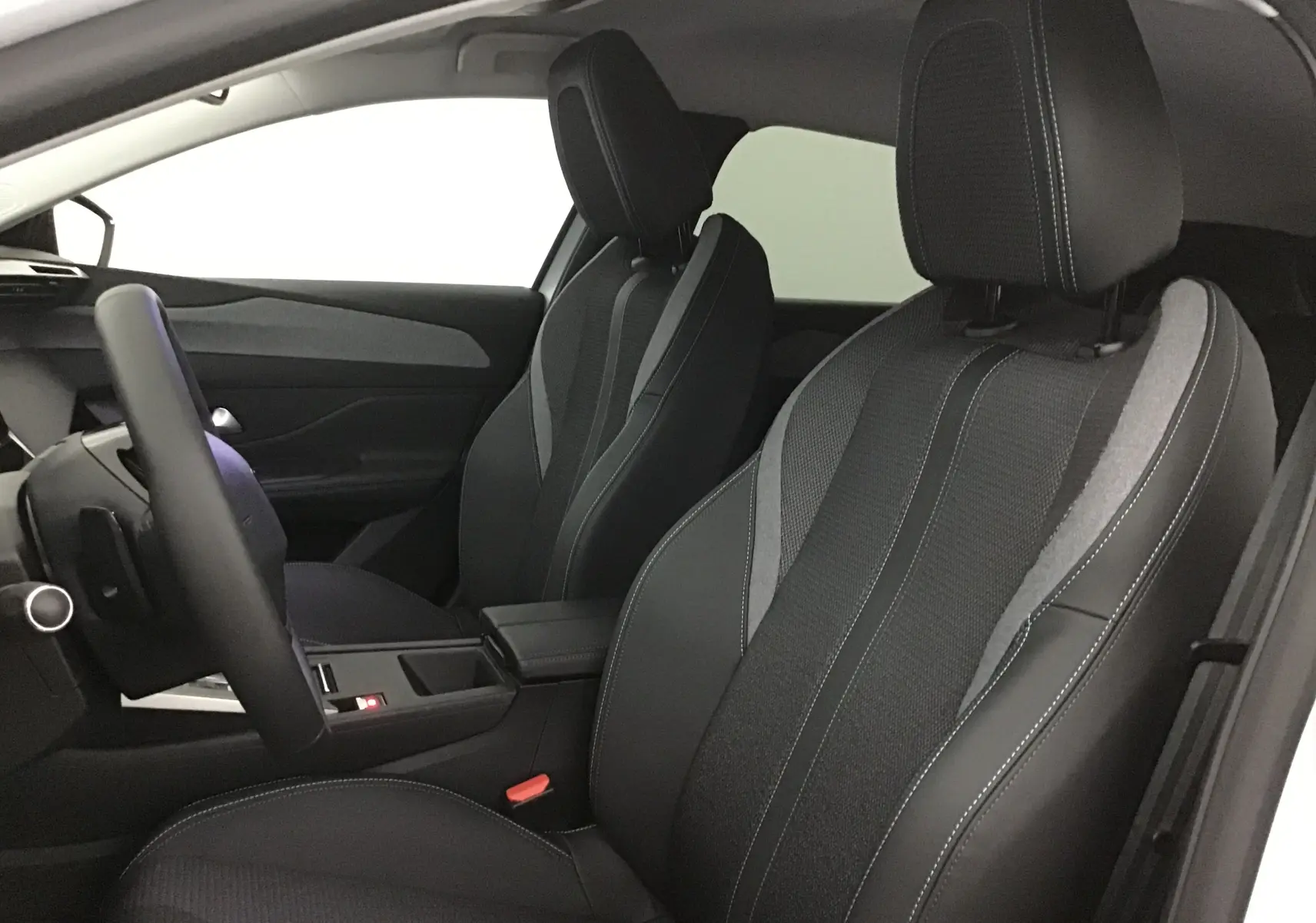 Intérieur de la Peugeot 308 2025, vue côté conducteur, sièges en tissu noir et gris avec surpiqûres blanches.