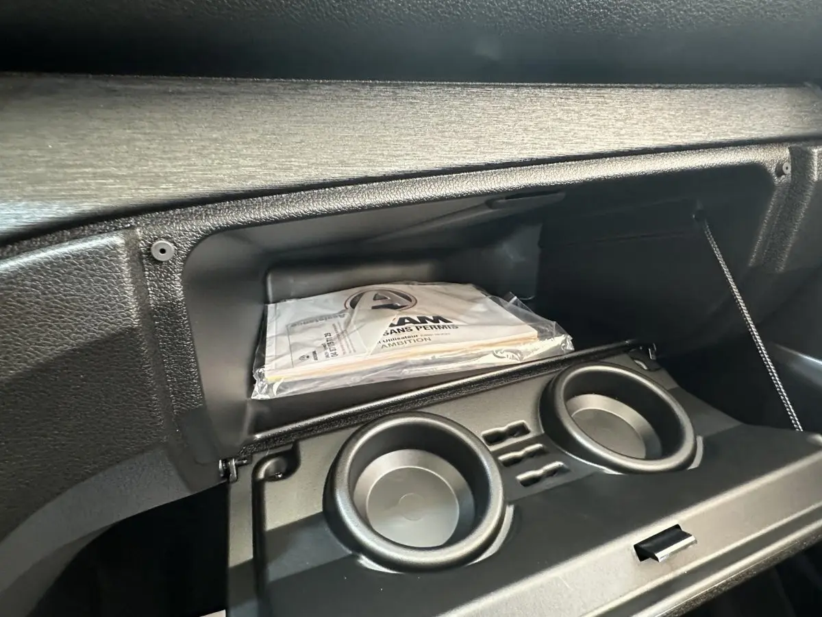 Intérieur de la boîte à gants ouverte de l'AIXAM AMBITION E CITY PACK blanc, avec porte-gobelets et manuel visible.