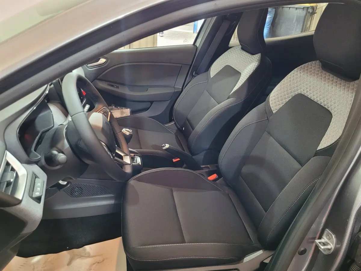 Vue intérieure côté conducteur de la Renault Clio gris foncé 2025, sièges tissu bicolore et volant multifonctions cuir synthétique.
