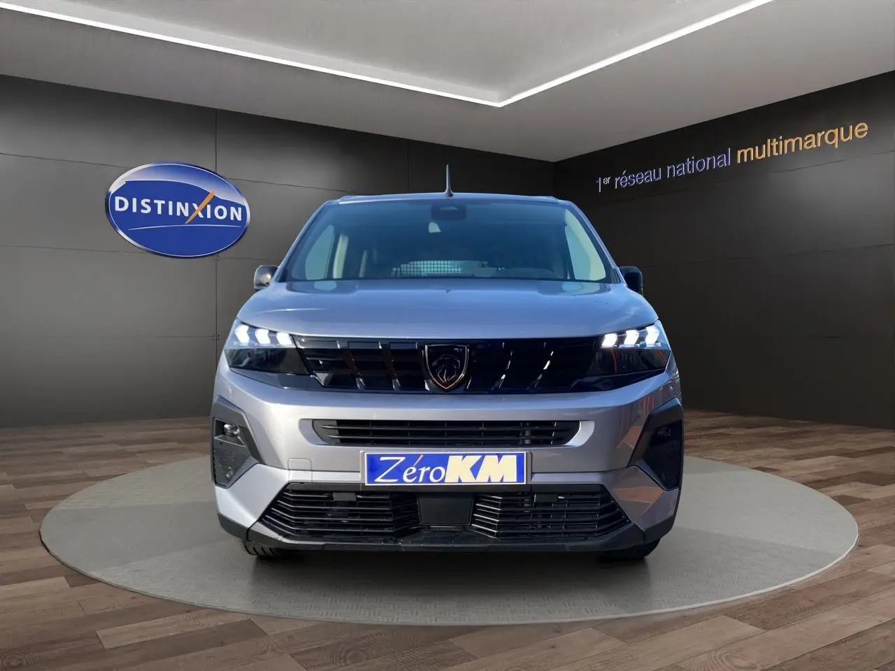Vue frontale d'un Peugeot Rifter gris acier 2026 avec calandre noire et phares LED allumés en showroom.