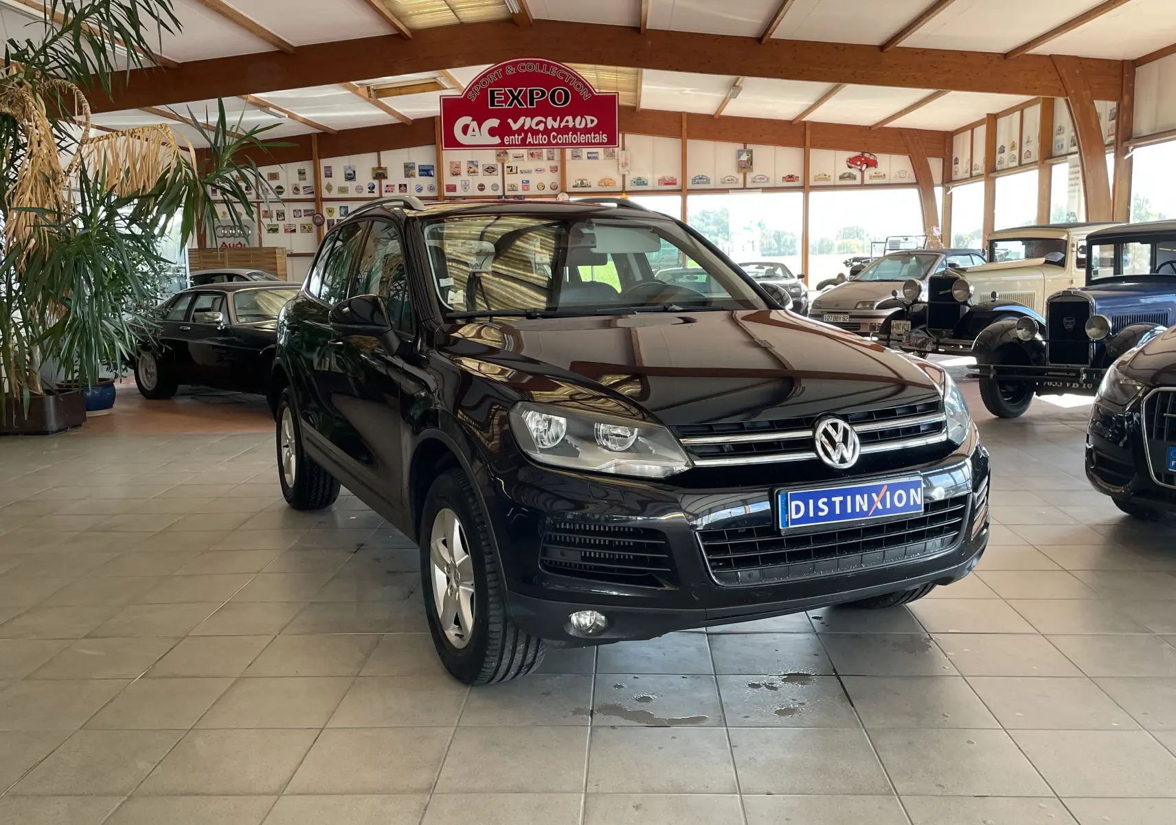 Volkswagen Touareg noir 3/4 avant droit dans un showroom avec plusieurs voitures anciennes en arrière-plan.