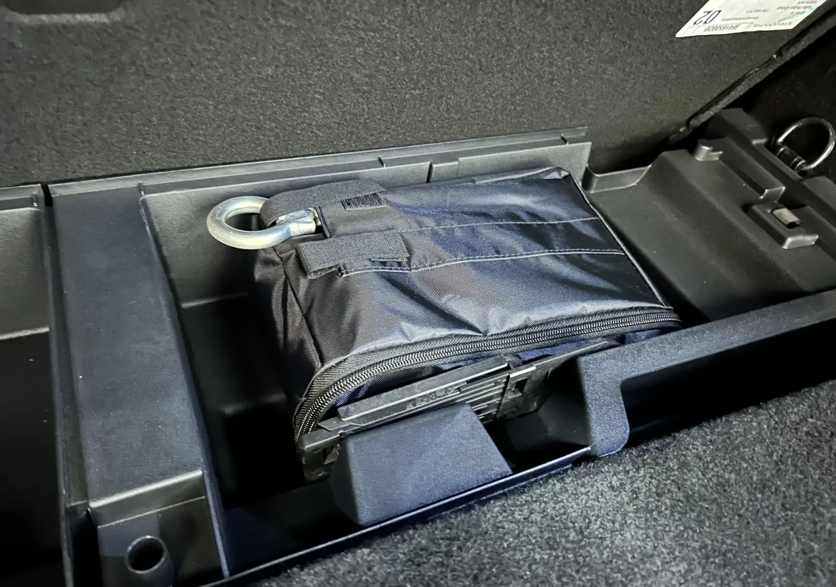 Rangement sous plancher de coffre d'une Peugeot 308 SW 2024, contenant une trousse noire avec anneau métallique.