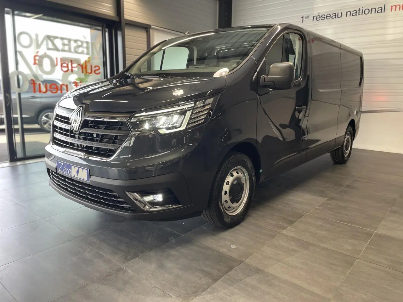 Vue 3/4 avant droit d'un Renault Trafic Fourgon gris comète avec projecteurs LED et jantes tôles 16 pouces en showroom.