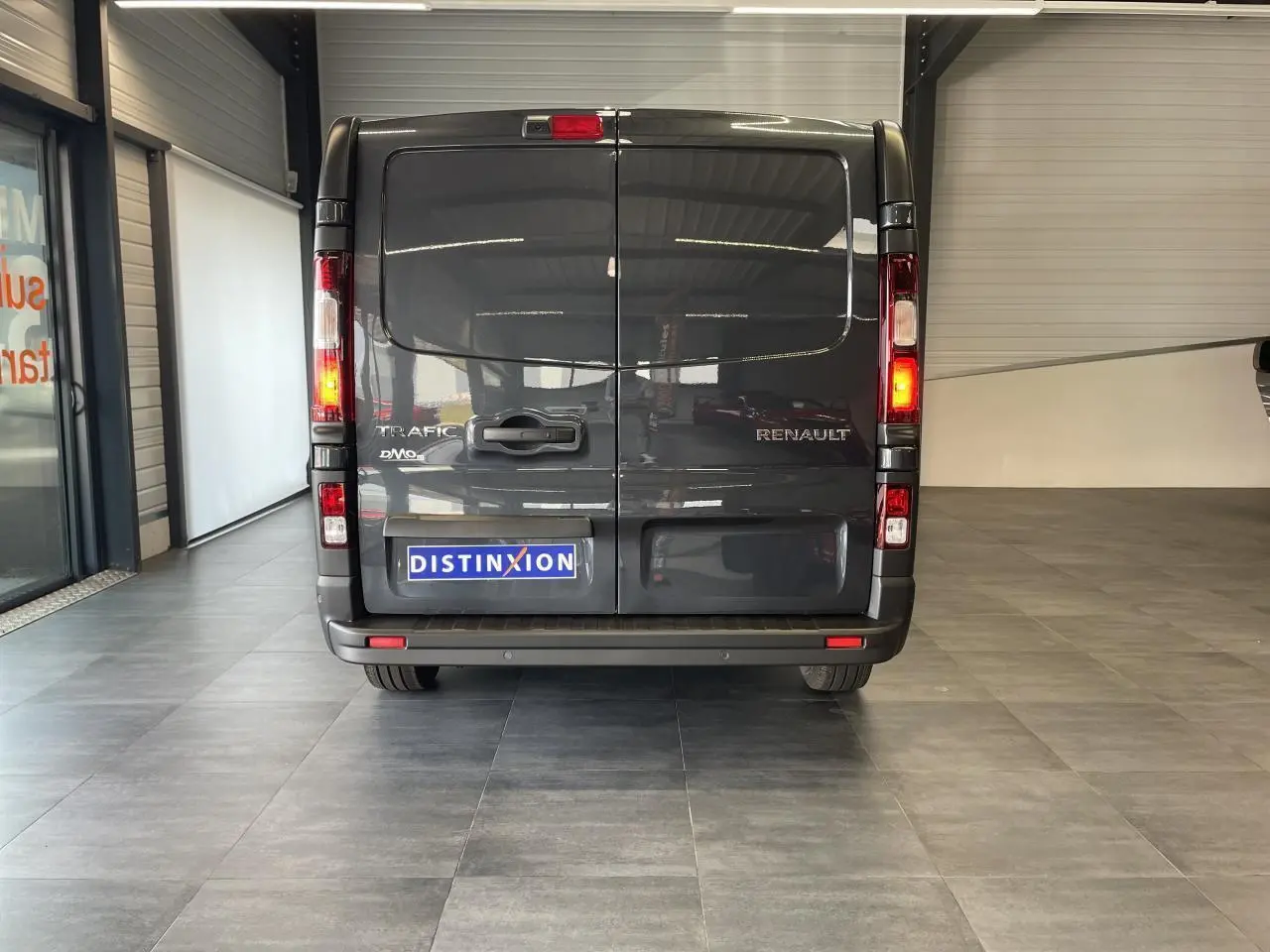 Vue arrière d'un Renault Trafic Fourgon gris comète avec feux allumés et plaque Distinxion visible en intérieur showroom.