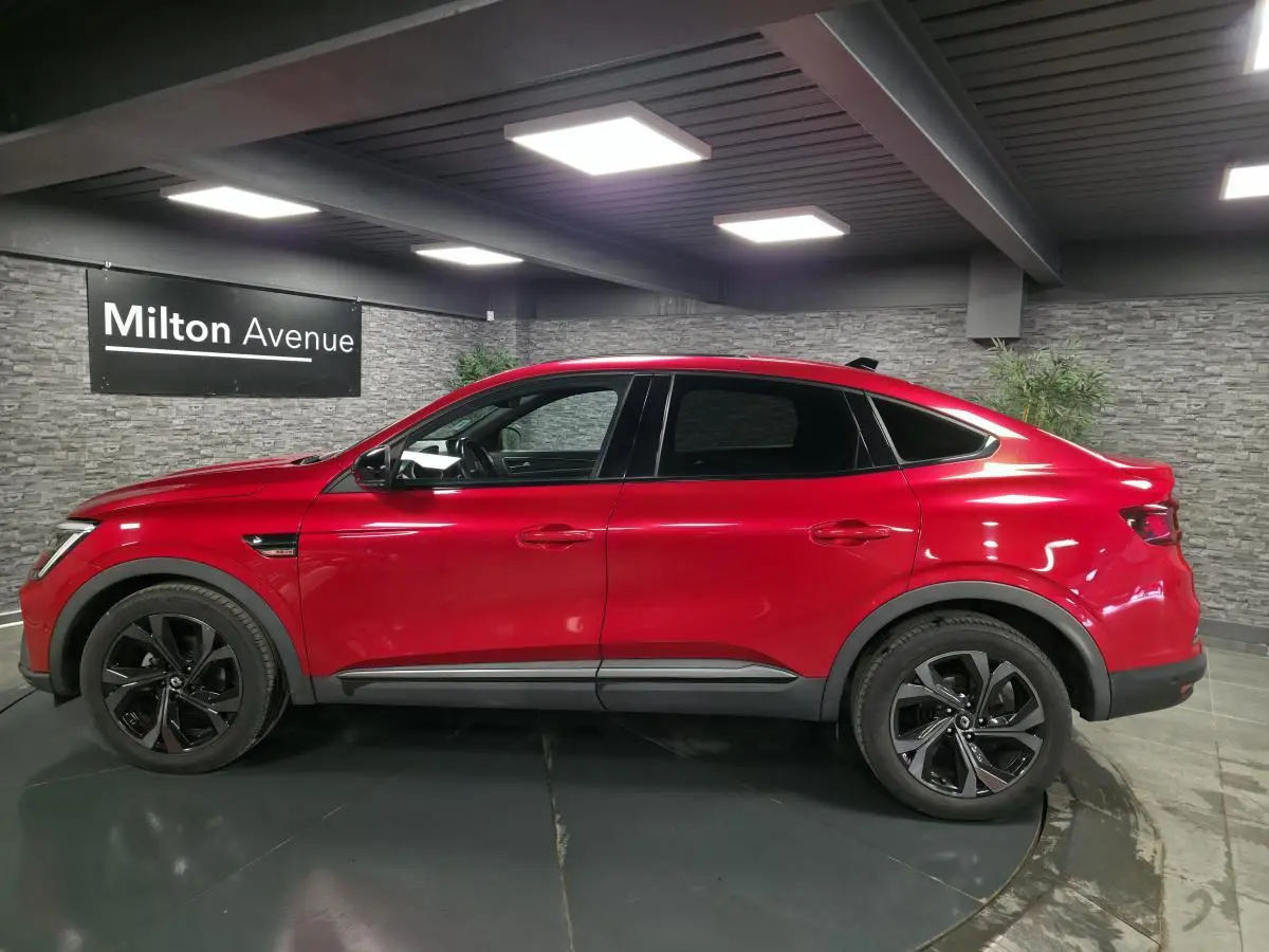 Profil droit du Renault Arkana 2023 rouge flamme avec jantes noires et toit ouvrant électrique visible en intérieur showroom.