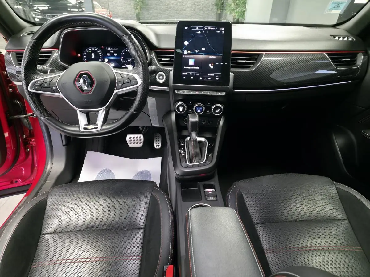 Vue intérieure avant du Renault Arkana rouge, tableau de bord avec écran tactile vertical et volant sport cuir noir à surpiqûres rouges.