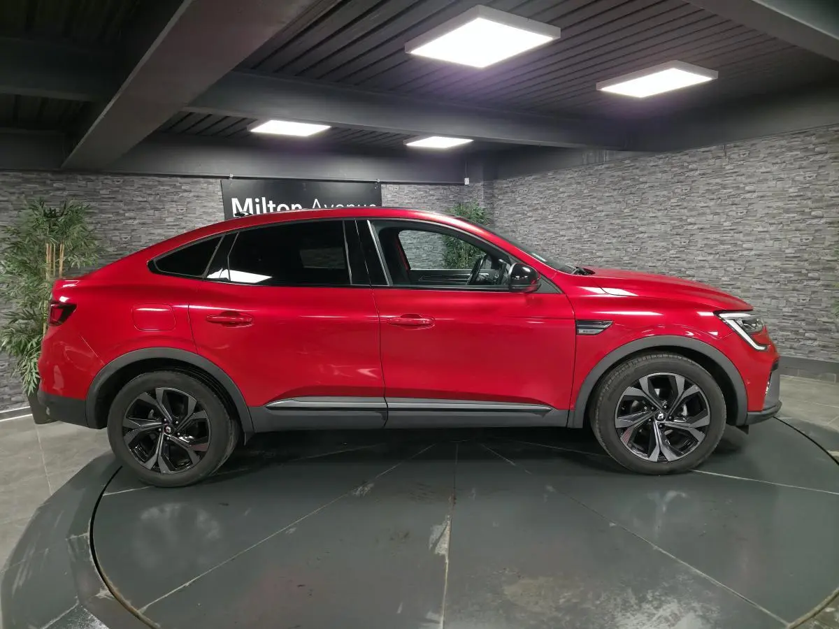 Profil droit d'une Renault Arkana rouge flamme 2023 avec jantes noires et toit ouvrant électrique visible en intérieur showroom.