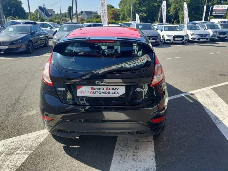 Vue arrière d'une Ford Fiesta noire avec toit rouge, stationnée dans un parking extérieur ensoleillé.