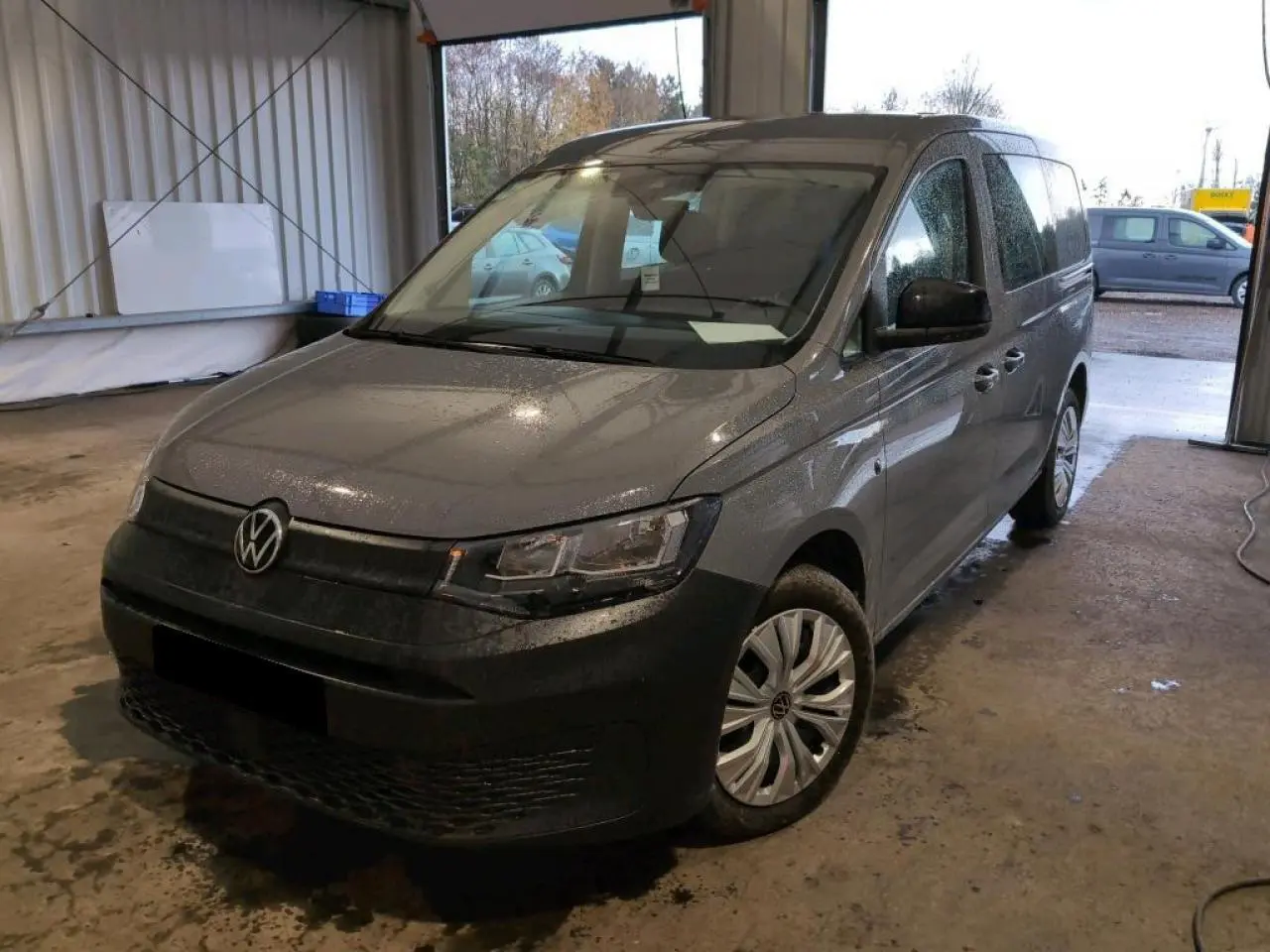 Vue 3/4 avant droit du Volkswagen Caddy Maxi gris Pure Grey, avec pare-chocs noir et jantes acier en intérieur d'atelier.