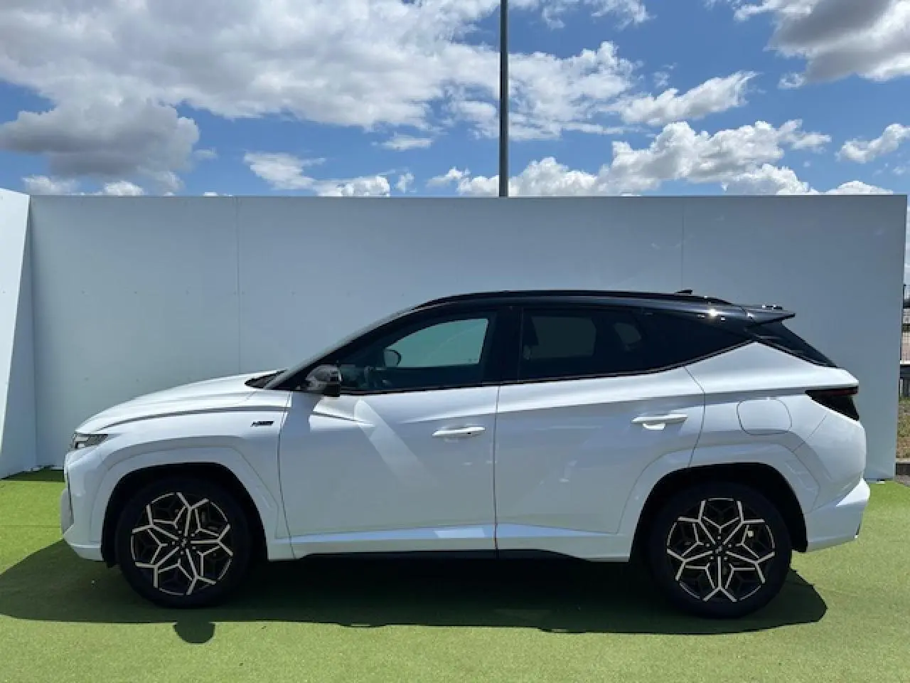 Profil côté gauche du Hyundai Tucson blanc polaire avec toit noir et jantes au design géométrique sous un ciel bleu.