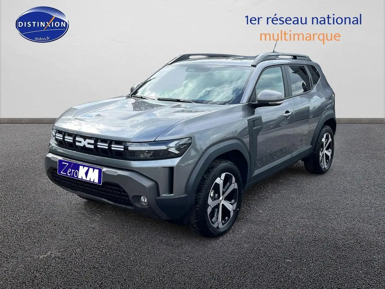 Dacia Duster gris schiste en 3/4 avant droit avec jantes alu et barres de toit visibles sur fond neutre.
