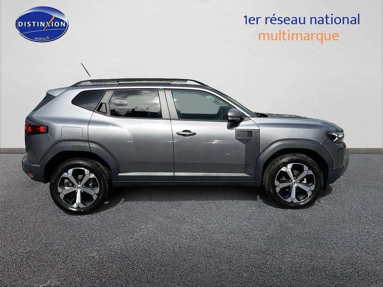 Profil droit du Dacia Duster gris schiste 2025 avec jantes alu et barres de toit visibles sur fond neutre.