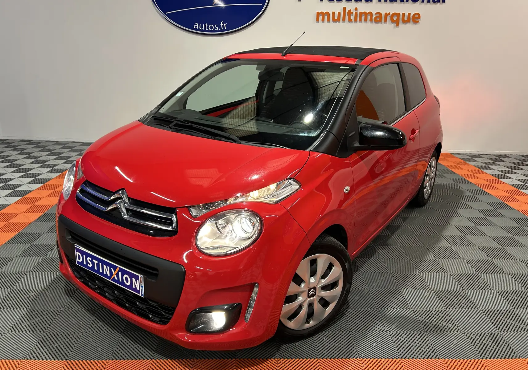 Citroën C1 rouge vue 3/4 avant droit, toit noir, feux allumés, dans un showroom avec sol gris et orange.