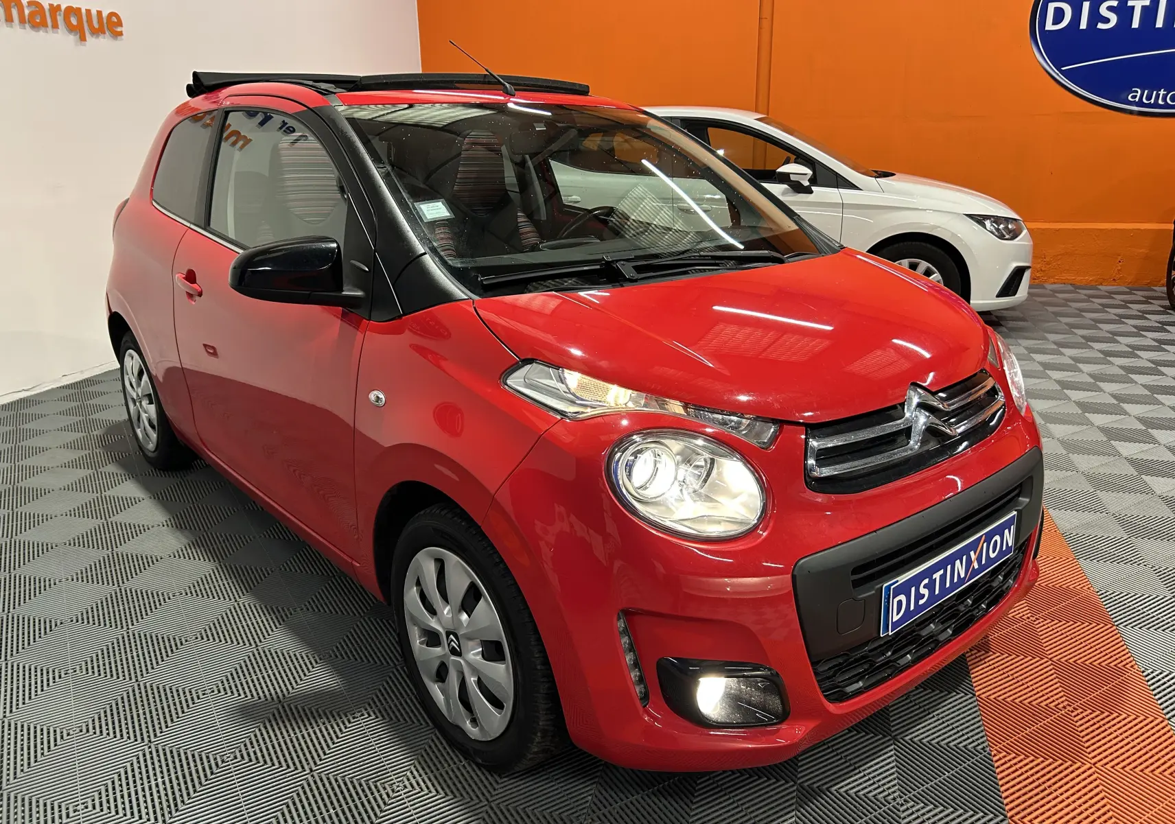 Citroën C1 rouge vue 3/4 avant droit avec toit ouvrant Airscape et phares allumés en intérieur showroom.