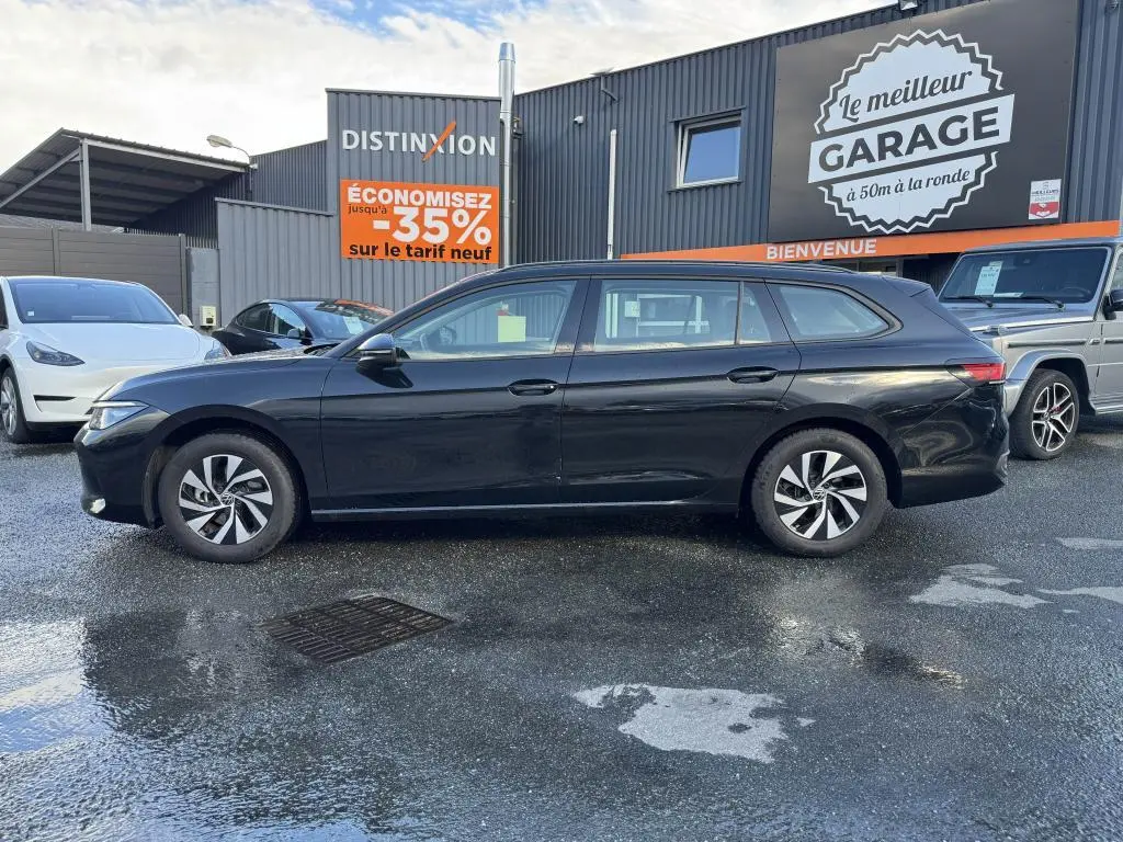 Volkswagen Passat SW noire vue de profil côté gauche, garée devant un garage avec panneaux publicitaires.