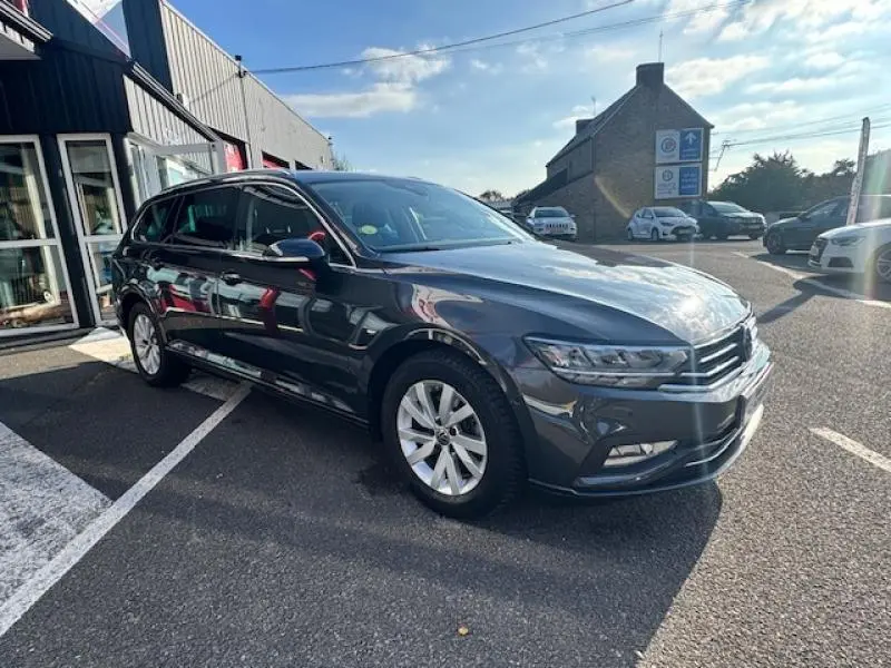 Volkswagen Passat SW gris manganèse métallisé vue 3/4 avant droit, avec jantes alu et barres de toit visibles.