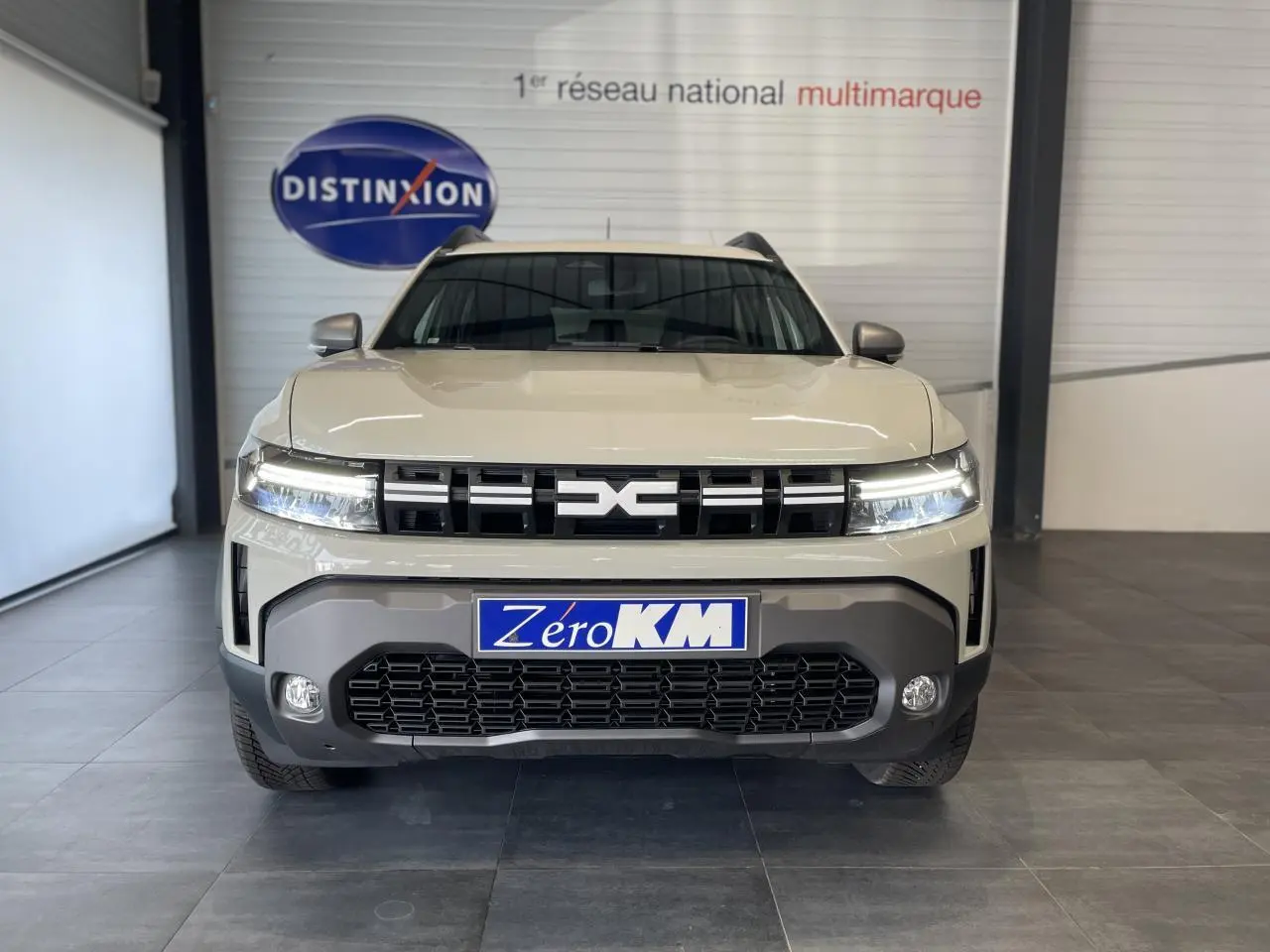 Vue frontale du SUV Dacia Duster beige Sandstone 2025 avec calandre noire et feux LED allumés en intérieur showroom.