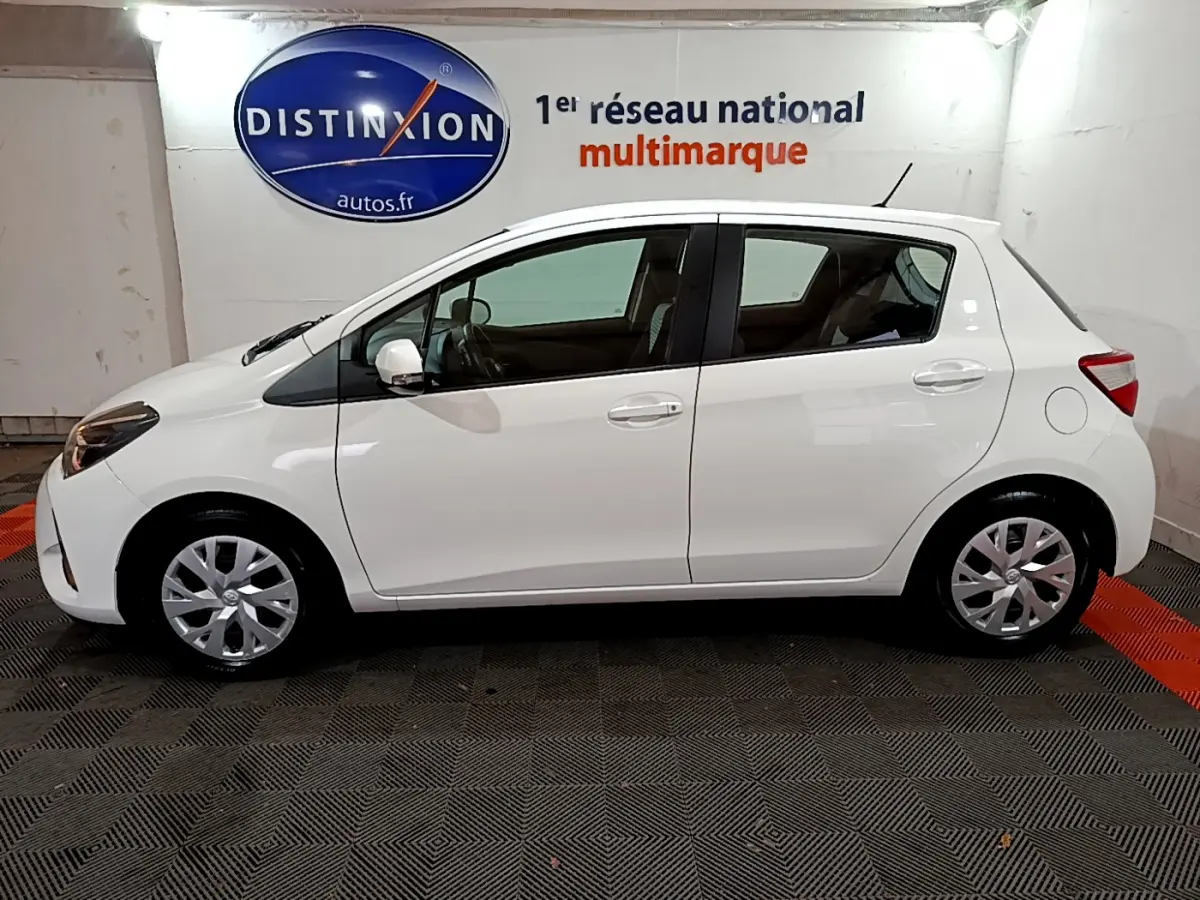 Vue de profil côté gauche d'une Toyota Yaris blanche 2020 hybride, stationnée en intérieur sur sol carrelé.