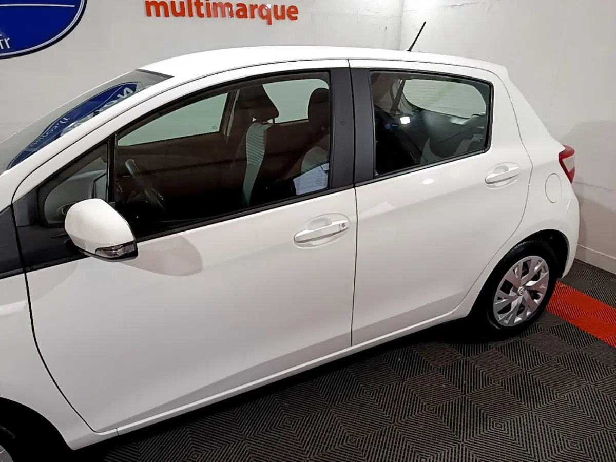 Vue de profil côté gauche d'une Toyota Yaris blanche 2020, version hybride, en intérieur sur sol à damier noir et rouge.