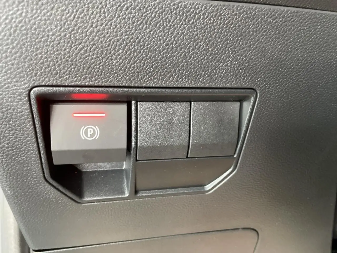 Gros plan sur le bouton de frein de stationnement électrique avec témoin rouge dans l'habitacle du Dacia Duster noir métallisé.
