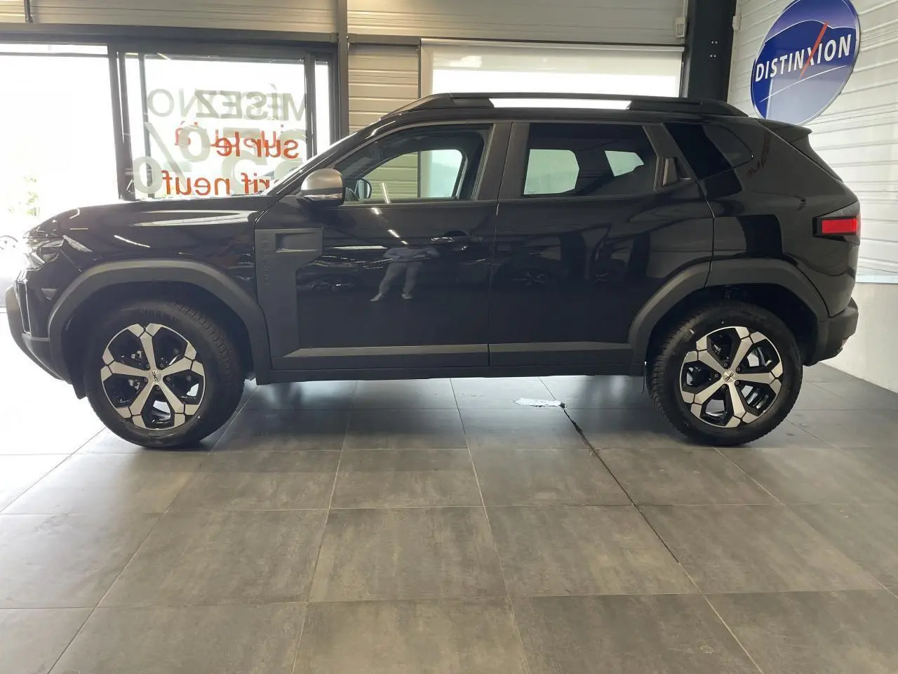 Profil côté gauche du Dacia Duster 2025 noir métallisé avec jantes alliage et barres de toit noires visibles en intérieur showroom.