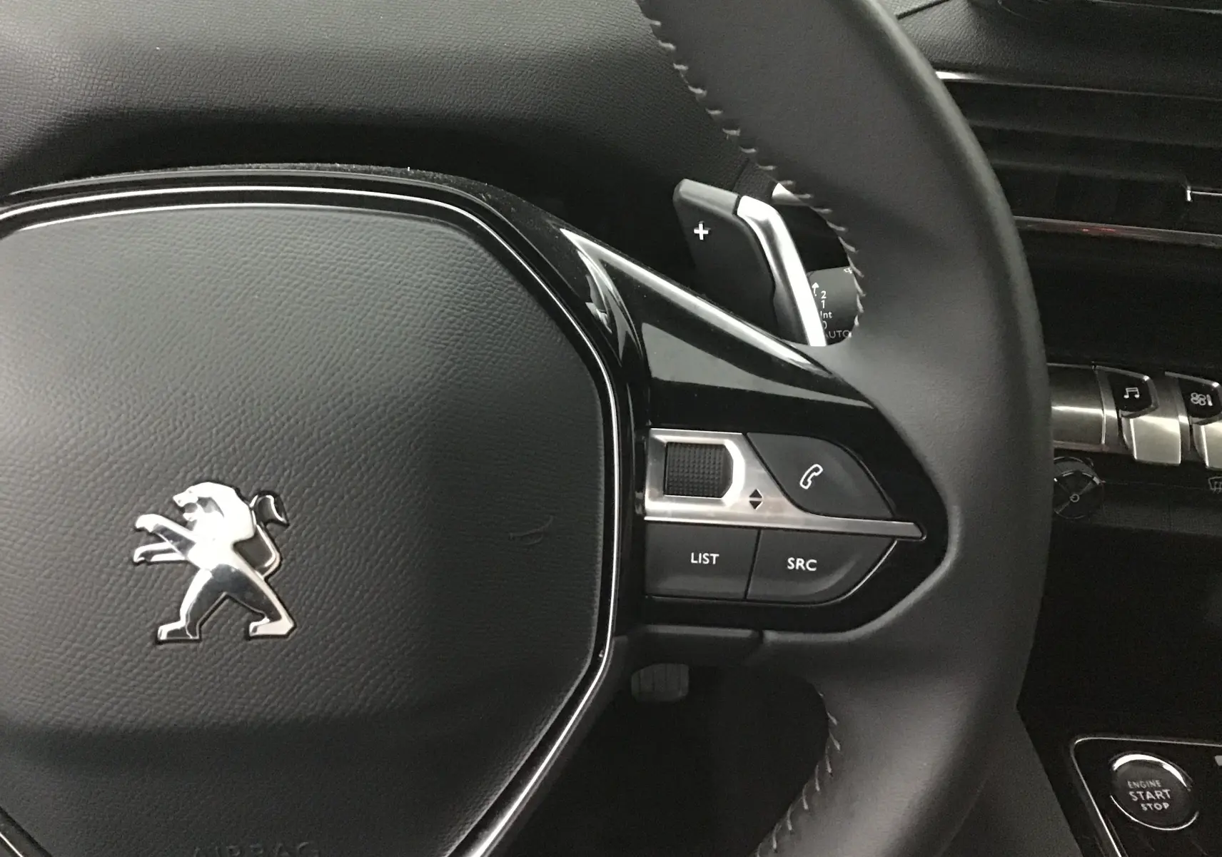 Gros plan sur le volant noir du Peugeot 5008 2024 avec commandes intégrées et logo lion chromé au centre.