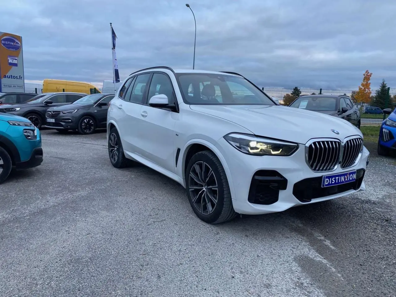 BMW X5 xDrive 30d blanc en 3/4 avant droit avec calandre M Sport et phares allumés sur parking extérieur.