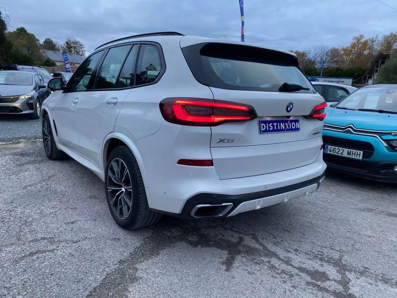 BMW X5 blanc vue 3/4 arrière droit avec feux allumés et double sortie d'échappement intégrée au pare-chocs.