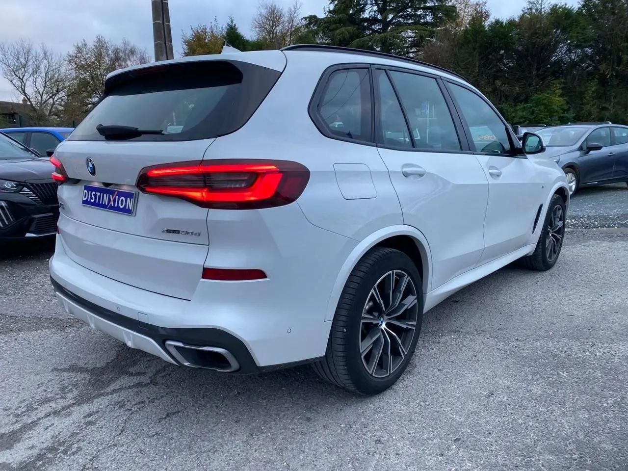 BMW X5 xDrive 30d blanc vu en 3/4 arrière droit avec jantes M Sport et feux arrière allumés.