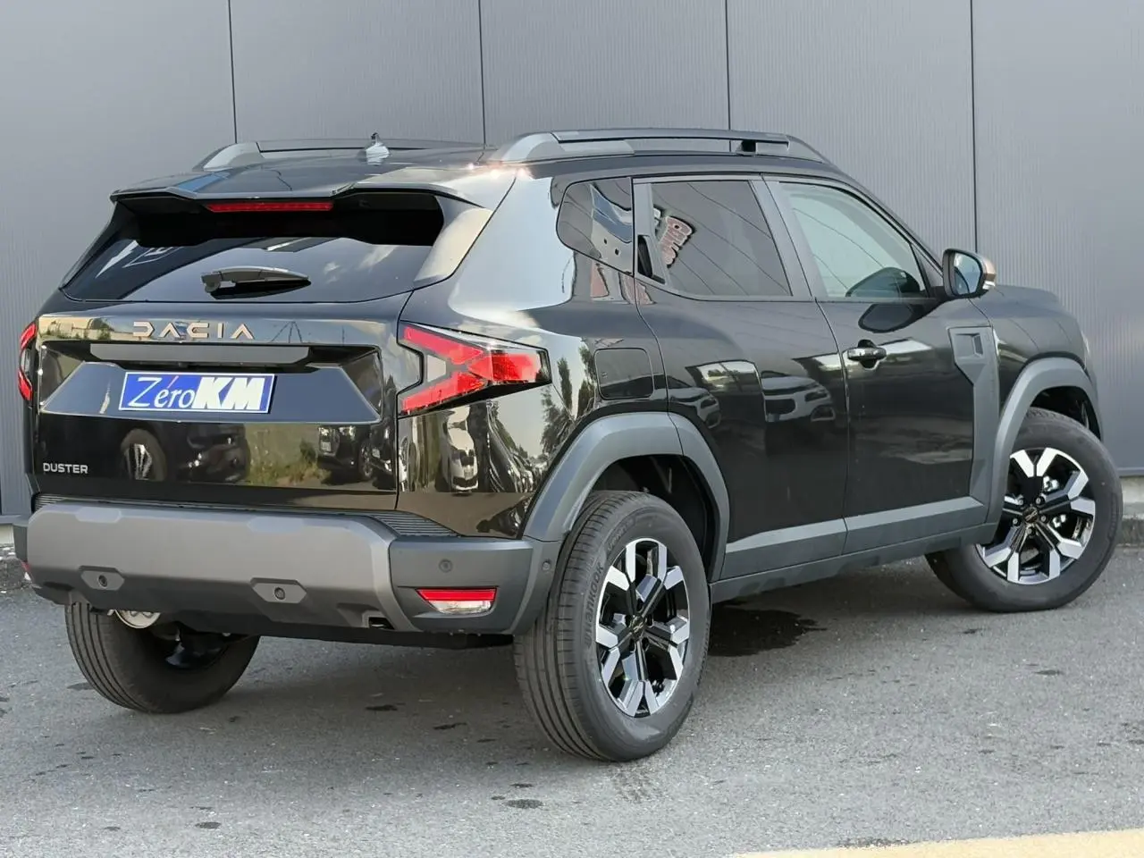 Vue 3/4 arrière droite du Dacia Duster noir nacré 2025 avec jantes alliage et barres de toit grises.
