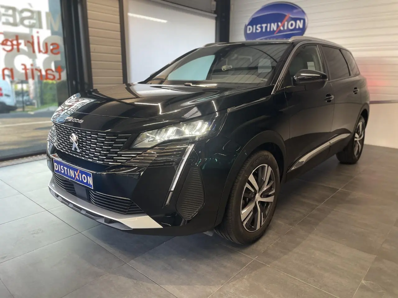 Peugeot 5008 noir métallisé vue 3/4 avant droit, avec calandre chromée et feux LED allumés en intérieur showroom.
