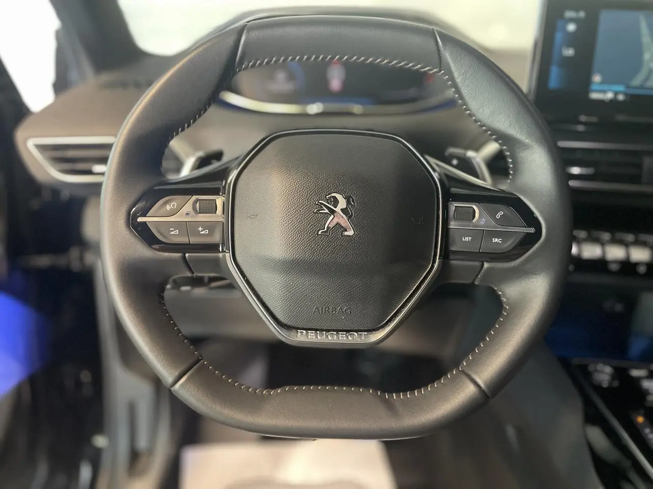 Volant compact en cuir noir avec logo Peugeot au centre, tableau de bord et écran tactile en arrière-plan flou.