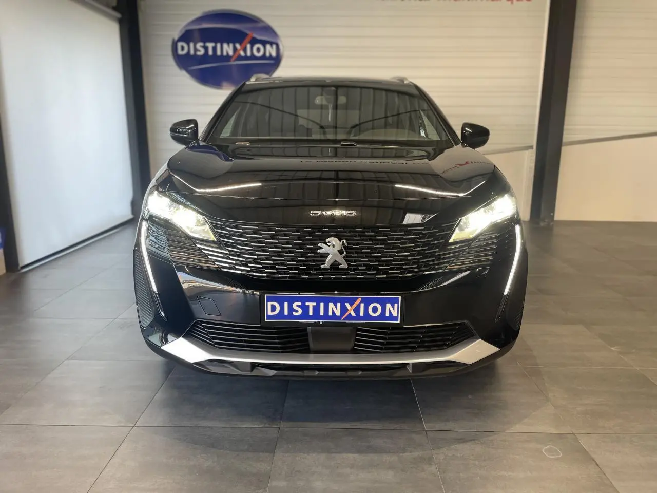 Vue frontale d'un Peugeot 5008 noir métallisé avec calandre à damier chromé et feux LED allumés en intérieur showroom.