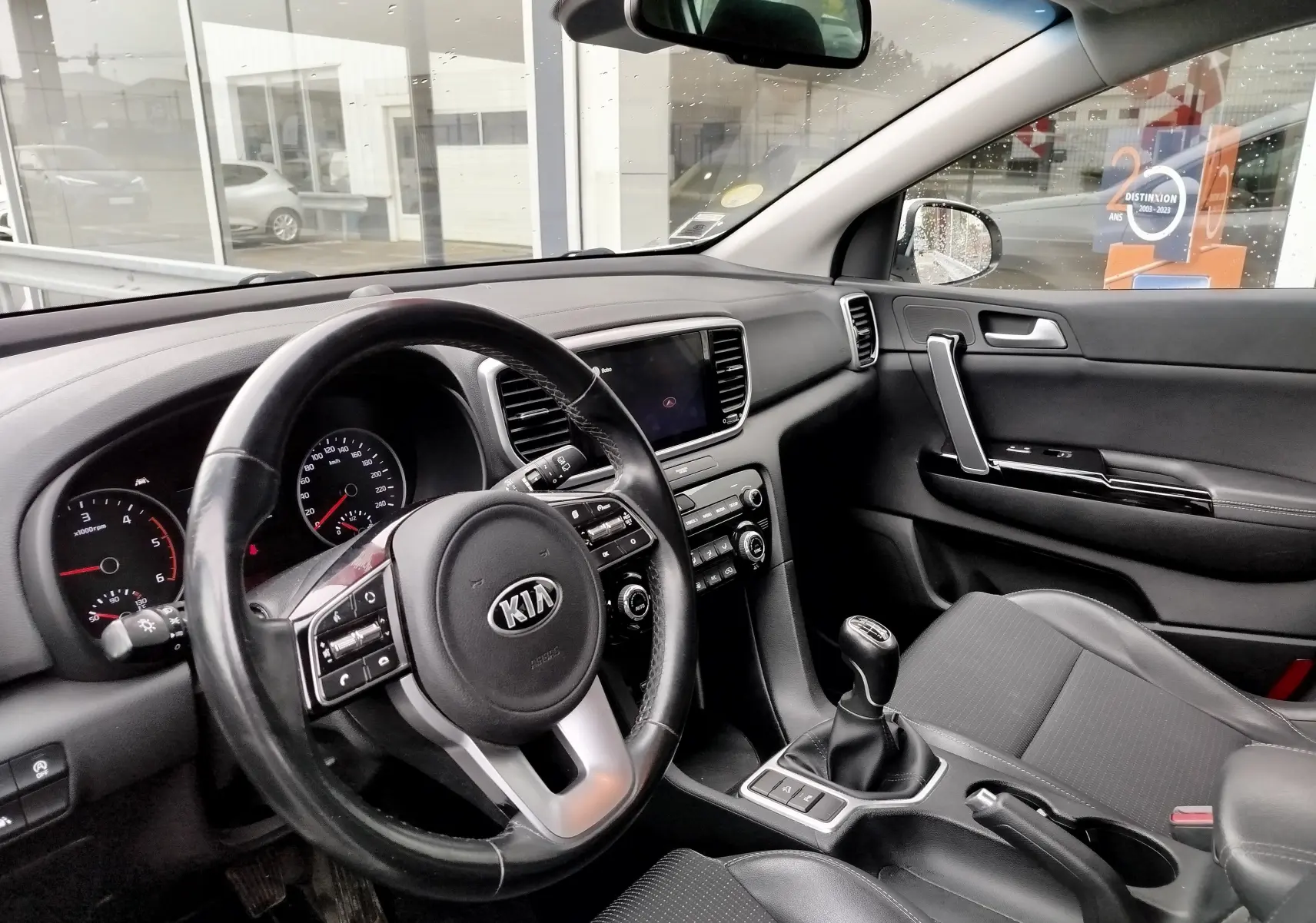 Intérieur du KIA SPORTAGE 2022 vu côté conducteur, volant cuir, tableau de bord noir et levier de vitesses manuel.