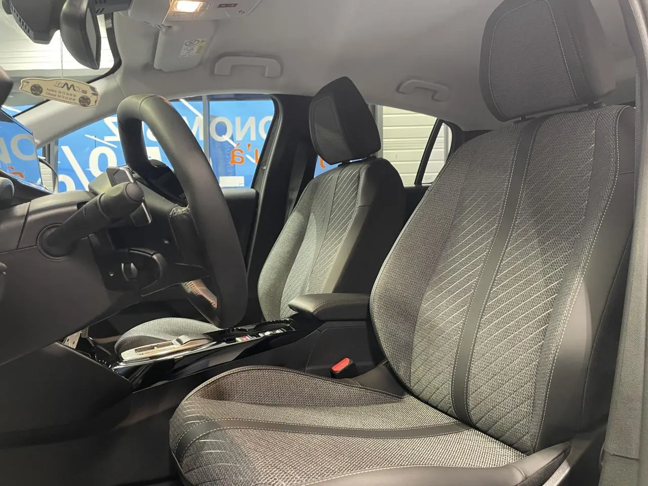 Vue intérieure côté conducteur de la Peugeot 208 1.2i Hybrid 2025 avec sièges tissu gris et accoudoir central noir.