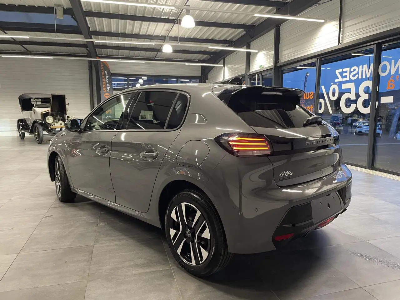 Vue 3/4 arrière droite d'une Peugeot 208 gris selenium avec feux arrière allumés et jantes alliage bi-tons en showroom.
