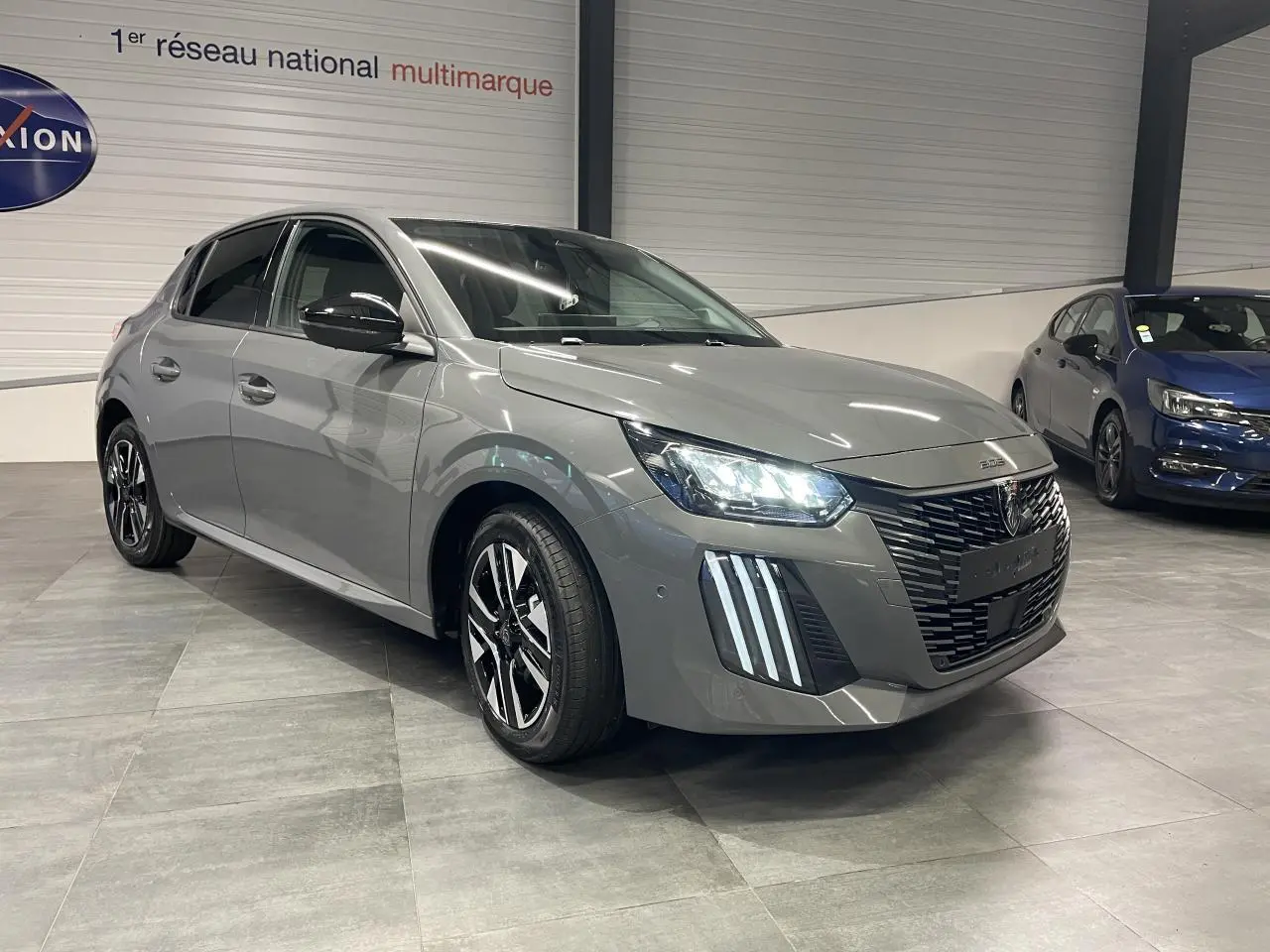 Peugeot 208 gris selenium en 3/4 avant droit avec calandre noire et feux LED allumés en intérieur showroom.