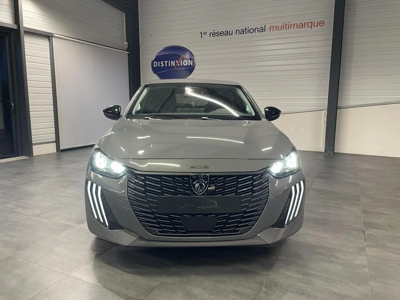Vue avant d'une Peugeot 208 gris sélénium avec calandre noire et feux diurnes LED en forme de griffes allumés dans un showroom.