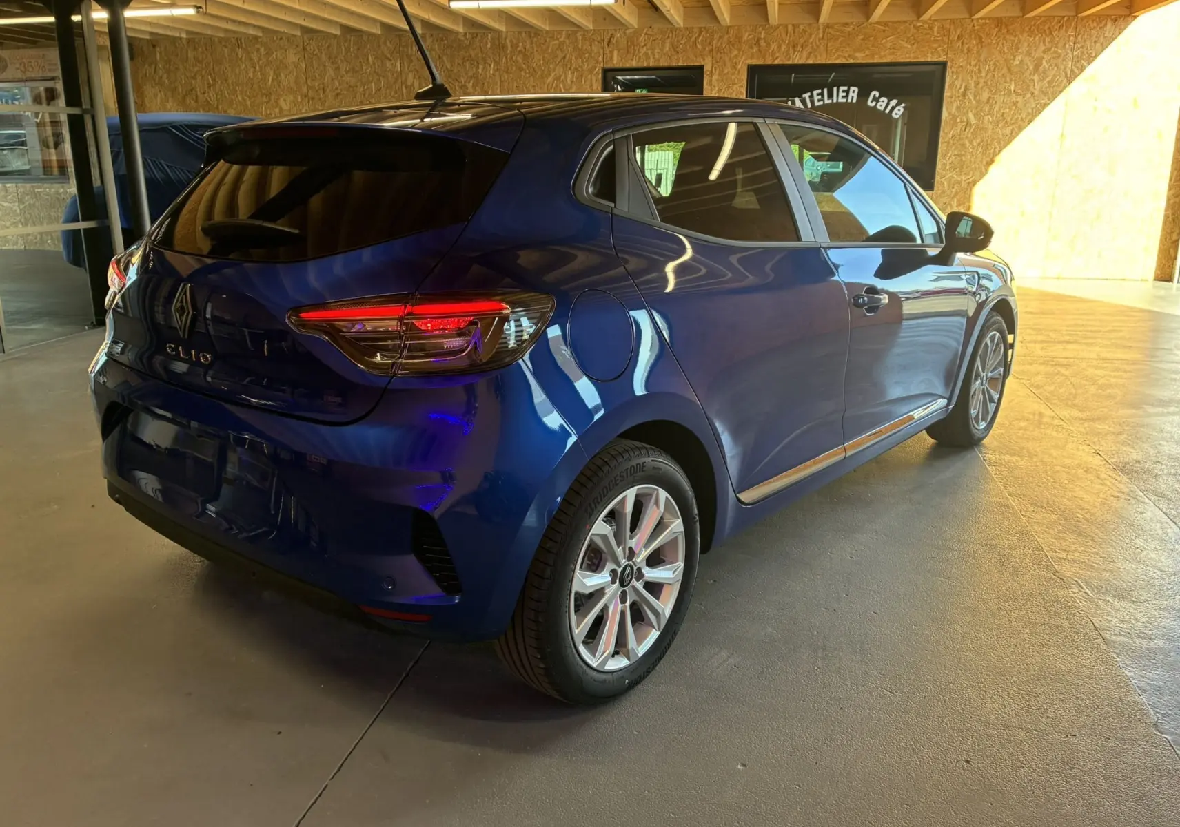 Vue 3/4 arrière droite d'une Renault Clio 2024 bleu iron, avec feux arrière allumés et jantes alliage visibles.