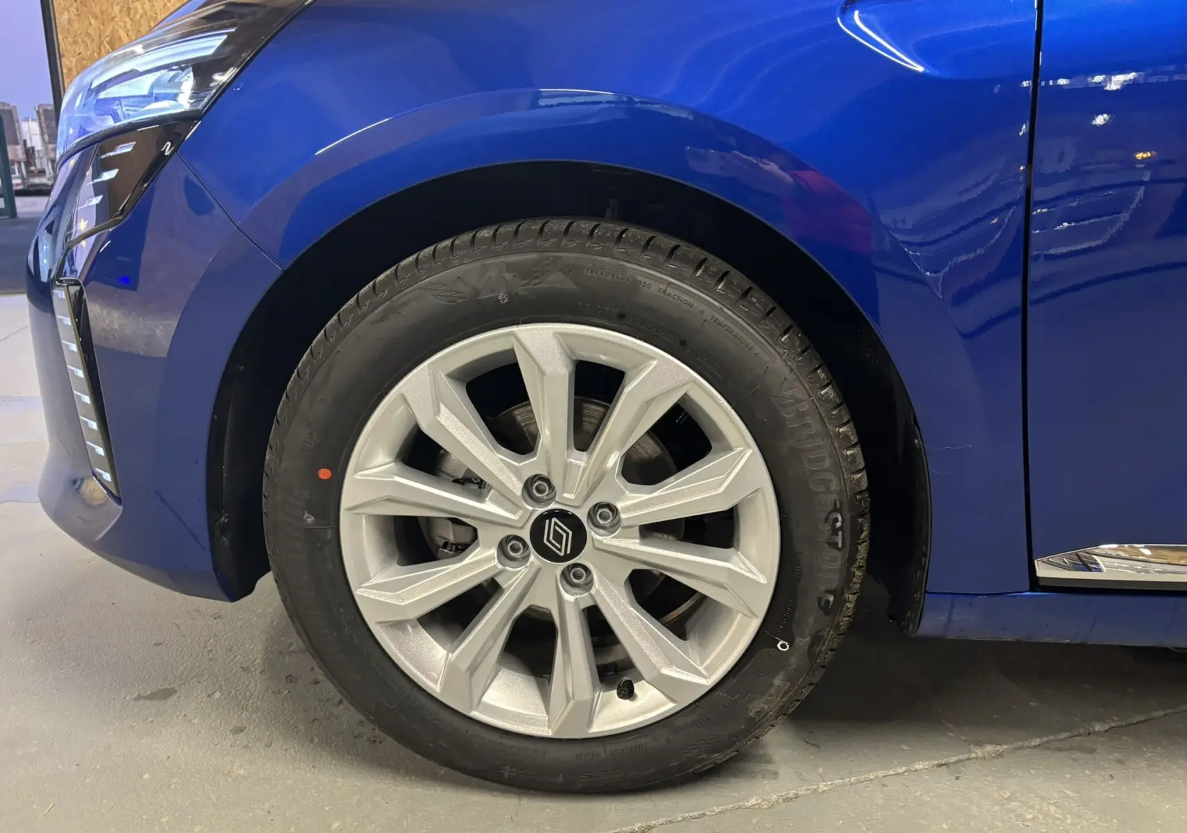 Gros plan sur la roue avant gauche d'une Renault Clio 2024 bleu iron avec jante alu et partie du pare-chocs avant visible.