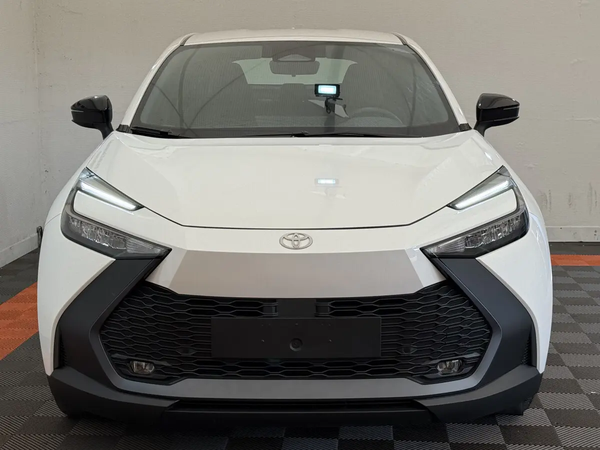 Vue frontale d'un Toyota C-HR 1.8 Hybride blanc avec calandre noire et phares LED fins, en intérieur sur sol damier.