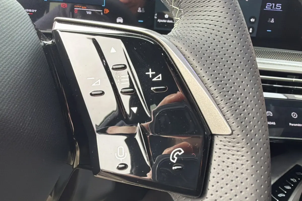 Gros plan sur les commandes gauche du volant cuir perforé du Peugeot 3008 Hybrid gris titane avec affichage numérique en arrière-plan.