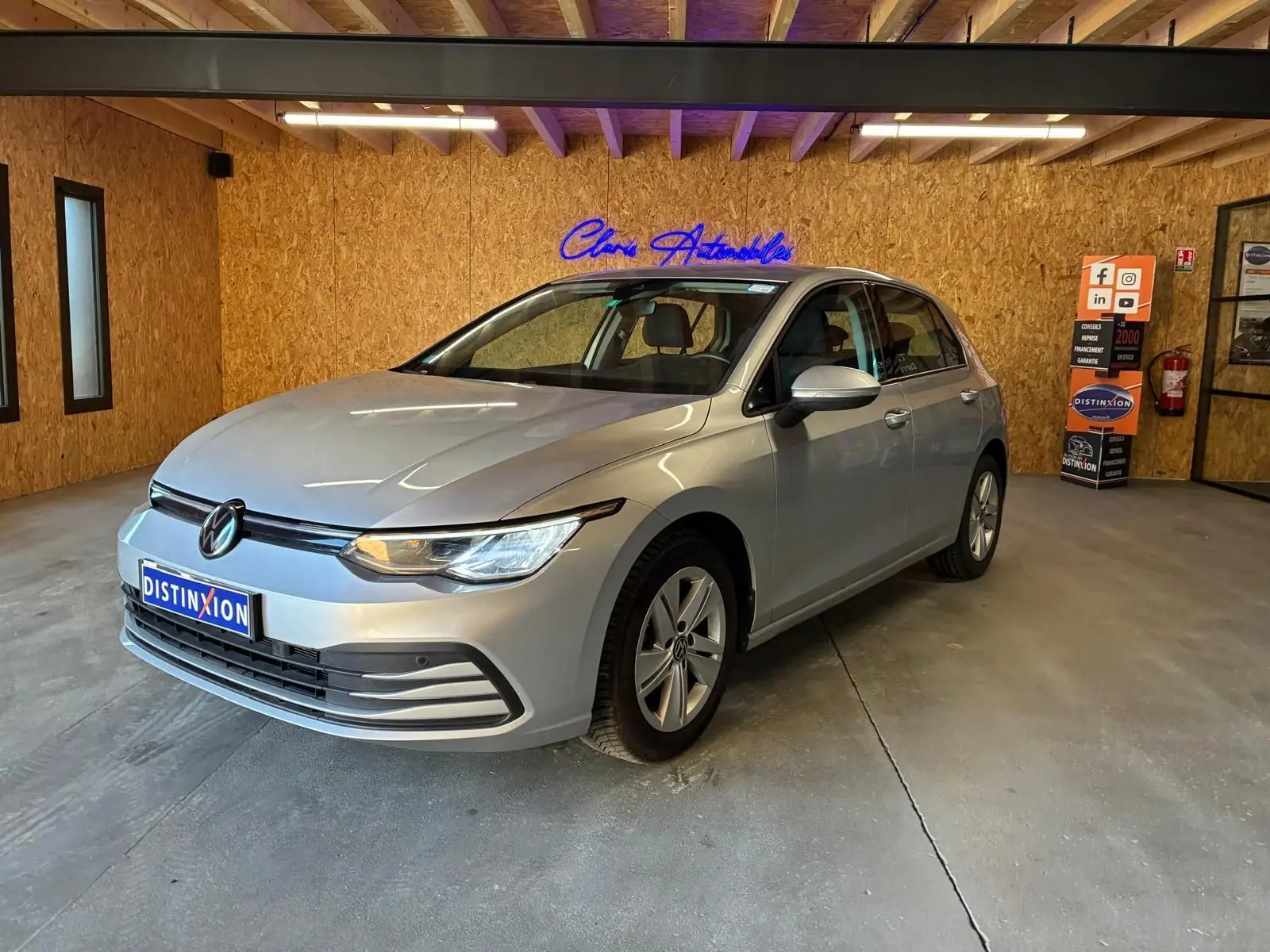 Volkswagen Golf gris clair vue 3/4 avant droit dans un showroom avec éclairage intérieur et panneau publicitaire.