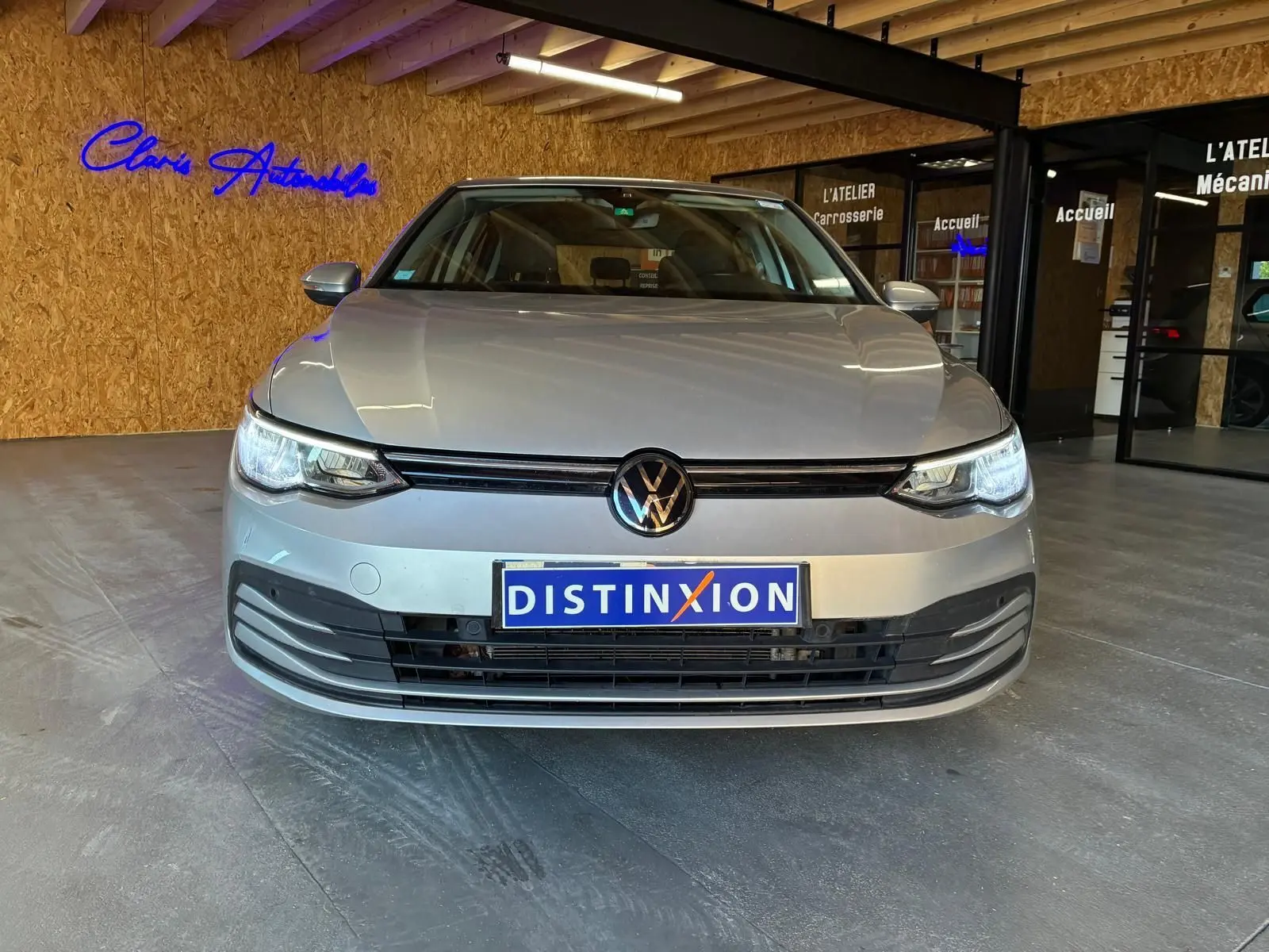 Vue avant d'une Volkswagen Golf 1.0 eTSI gris clair 2023 avec plaques Distinxion dans un showroom.