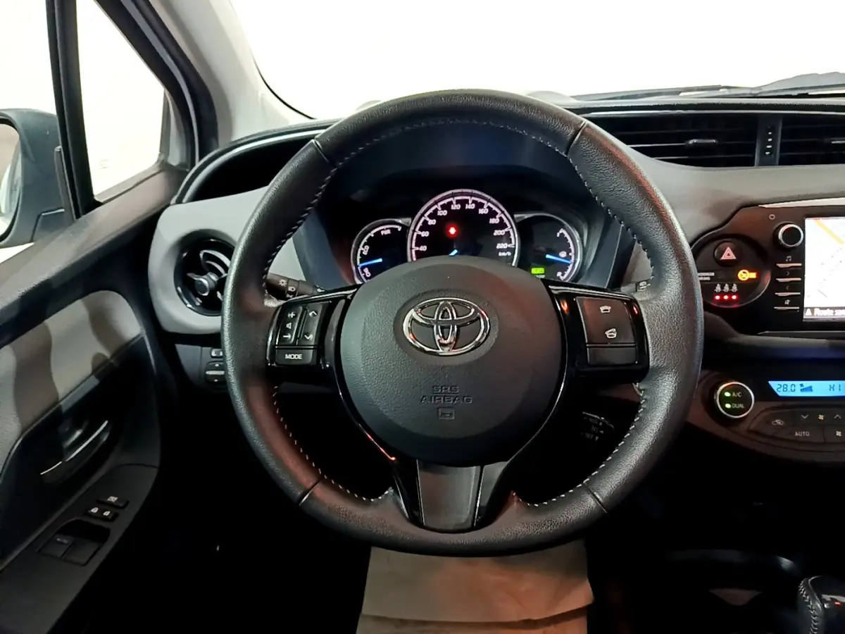 Vue intérieure centrée sur le volant cuir et alu de la Toyota Yaris blanche, avec tableau de bord et écran tactile visibles.