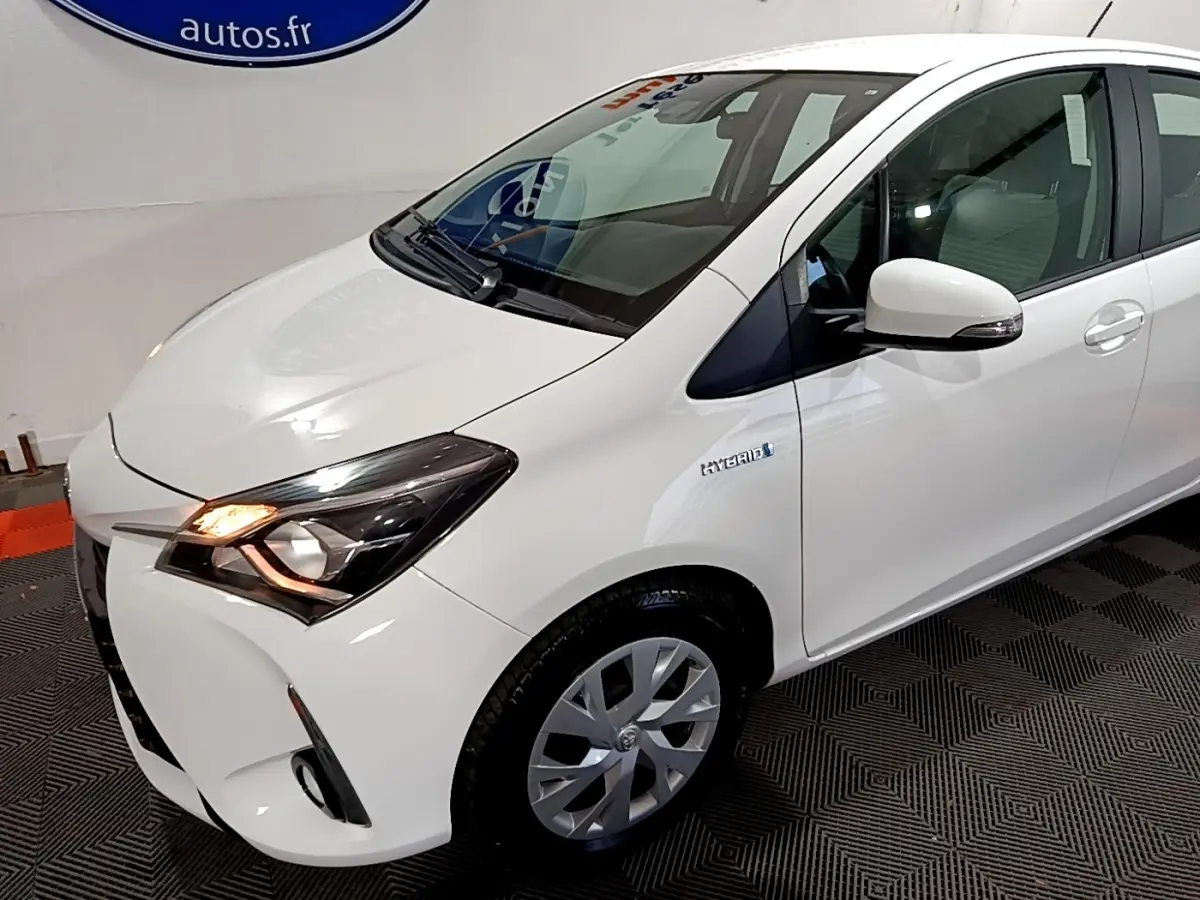 Vue 3/4 avant droit d'une Toyota Yaris blanche hybride avec phares allumés et badge Hybrid sur l'aile avant.