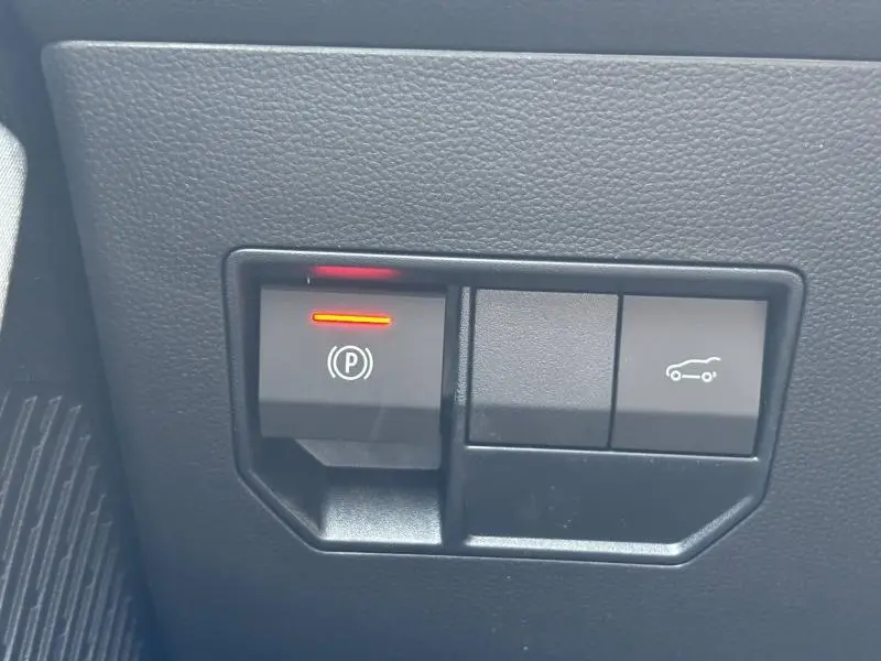 Gros plan sur les commandes noires du frein à main électronique et de l'ouverture du coffre dans un Dacia Bigster blanc.