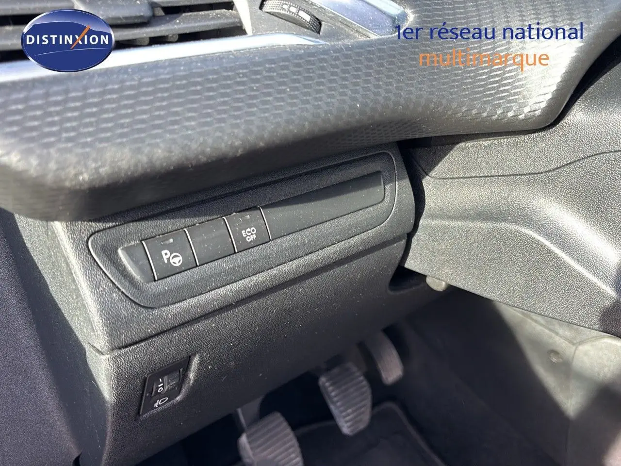 Détail des commandes côté conducteur du tableau de bord noir avec pédales d’embrayage, frein et accélérateur visibles.