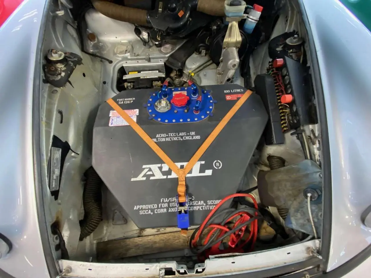 Vue plongeante sur le compartiment avant gris métallisé d'une Porsche 911 3.0 SC avec réservoir FIA noir et sangles orange visibles.