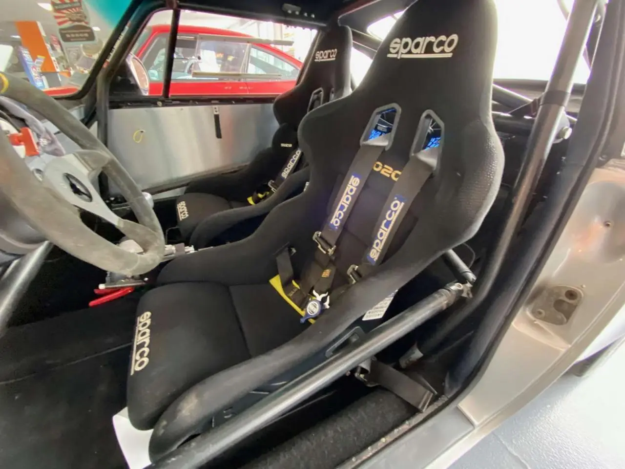Intérieur de Porsche 911 3.0 SC gris, vue côté gauche sur baquets Sparco noirs avec harnais et arceau tubulaire.